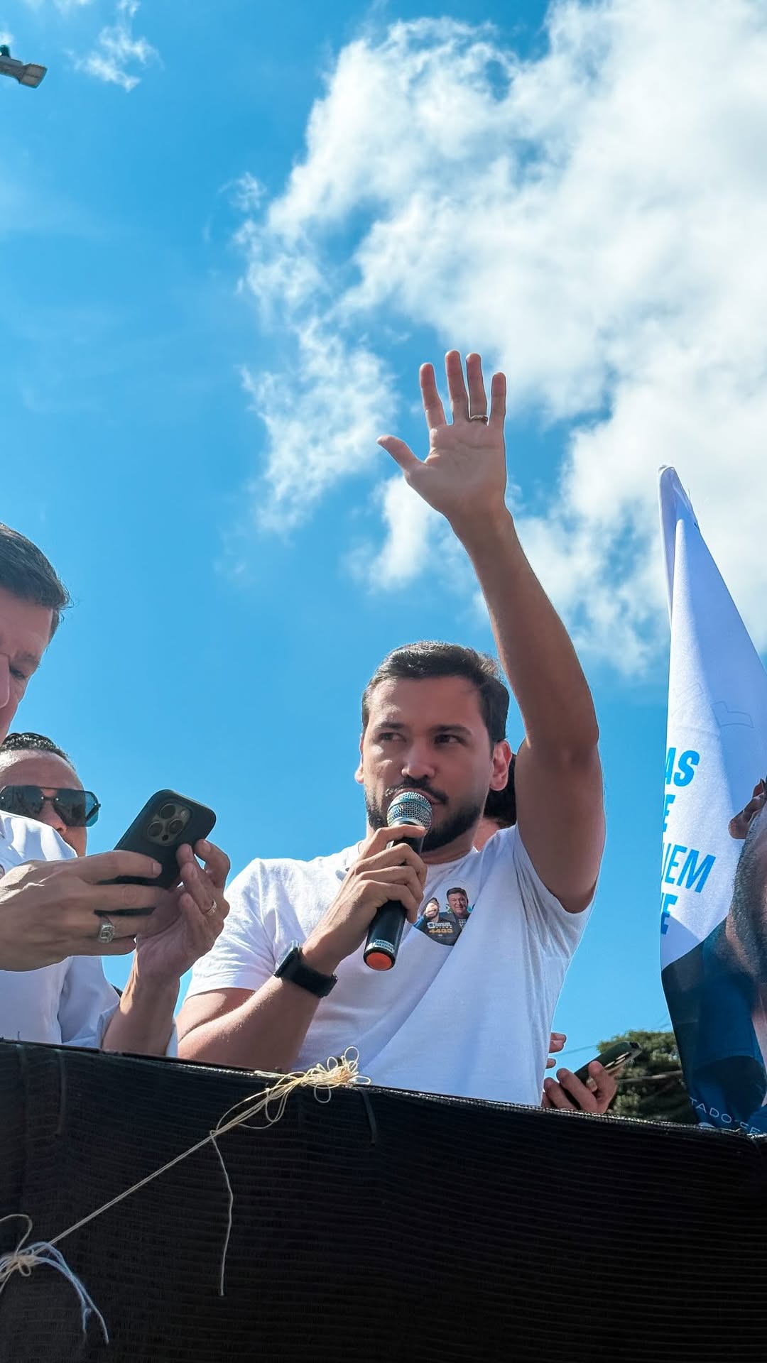 Samuel Viana discursa ao microfone com a mão erguida durante ato de campanha ao ar livre em Minas Gerais, ao lado de apoiadores e de bandeira com o slogan Minas segue com quem resolve.